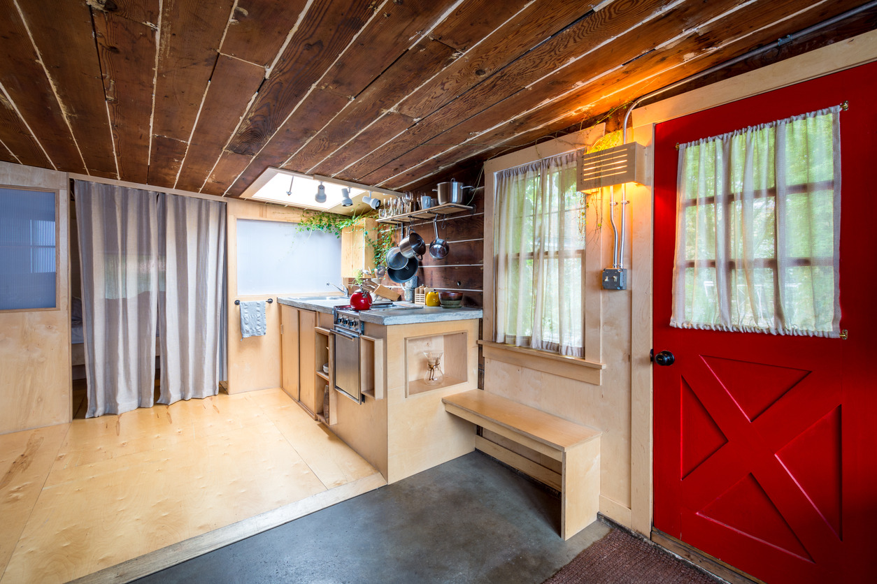 This charming interior showcases a cozy home environment featuring rustic wood finishes, a bright red door, and simple, functional kitchen space. The wood-paneled ceiling and walls create a warm, inviting atmosphere, complemented by natural light filtering through curtained windows.