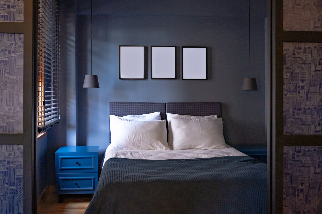 A stylish and inviting bedroom features a comfortable bed with white pillows and a gray patterned headboard. The walls are painted in a dark blue-gray tone, complemented by matching curtains and a striking blue nightstand.